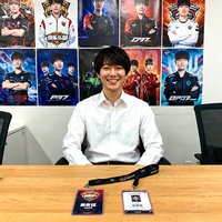 一位穿着白色衬衫的俱乐部运营人员，坐在贴满战队海报的办公室内，微笑面对镜头，桌上摆放着赛事通行证和工作牌